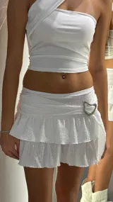 Minifalda blanca de tiro bajo con diseño de volados superpuestos y textura arrugada. Presenta una hebilla metálica decorativa en forma de corazón en la cintura.