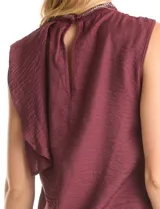 Blusa sin mangas color blanco, confeccionada en crepe con acabado satinado. Presenta un cuello alto decorado con mostacillas y una sobrecapa drapeada en el frente. La espalda cuenta con una abertura tipo gota y cierre de botón.