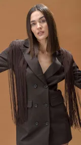 Blazer tipo vestido color chocolate, de corte cruzado con doble botonadura frontal. Presenta solapas clásicas y flecos largos decorativos en los hombros y mangas que aportan movimiento.