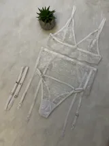Conjunto de lencería de encaje blanco compuesto por un bralette triangular, una tanga con tiras y un portaligas integrado. El diseño presenta detalles de encaje floral y breteles regulables.