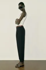 Pantalón tobillero de tiro medio, color beige, con pinzas delanteras y bolsillos frontales. Incluye un cinturón fino de cuero marrón con hebilla metálica dorada y cierre frontal con cremallera, botón interior y gancho.
