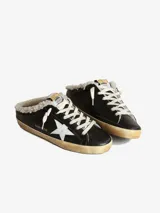 Zuecos Golden Goose modelo Super-Star Sabot, con diseño de zapatilla sin talón. Presentan exterior cubierto de glitter negro y dorado, estrella característica en el lateral, cordones y forro interior de shearling sintético.