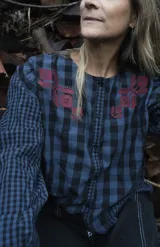 Blusa de algodón con estampado a cuadros en tonos azul y negro, con bordados decorativos en color bordó sobre el canesú. Presenta cierre frontal con botones, cuello redondo y mangas largas levemente abullonadas.