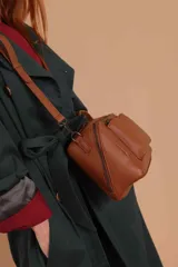 Bandolera de diseño rectangular en color suela, confeccionada en material sintético. Cuenta con un asa superior fija, correa larga ajustable y extraíble, y dos bolsillos frontales con cierre metálico, uno de ellos con diseño superpuesto. Presenta el logo de la marca grabado en el frente.