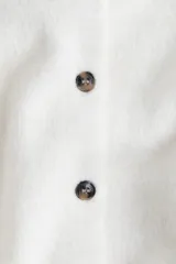 Cárdigan corto de punto suave color crema, con cuello camisero, abotonadura frontal con botones marrones y remate acanalado en puños y bajo.