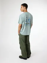 Remera oversize verde menta de manga corta y cuello redondo, confeccionada en algodón orgánico. Presenta un estampado gráfico en la espalda con diseño de surfistas y texto en caracteres japoneses y latinos.