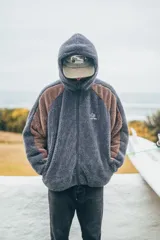 Campera con capucha de tejido sherpa, combinando paneles en color gris y marrón. Presenta cierre frontal completo, mangas largas y diseño holgado.