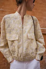 Chaqueta tipo bomber de cuello redondo y manga larga, confeccionada en tejido jacquard con diseño floral en tonos dorados y crema. Presenta cierre frontal con cremallera, bolsillos delanteros de plastrón con solapa y puños y bajo elásticos.