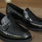 Mocasín de cuero vacuno negro con textura grabada tipo reptil. Presenta un diseño clásico con antifaz en el empeine, suela de goma en tono natural y taco bajo de madera.