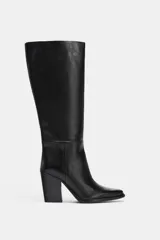 Bota alta de cuero negro, estilo cowboy, con tacón de bloque alto y punta cuadrada. Presenta costuras decorativas en el cuerpo de la caña.