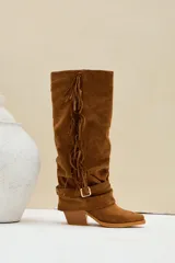 Botas altas estilo texana de gamuza color camel, con taco cuadrado y puntera fina. Presentan flecos largos colgando a lo largo del lateral de la caña y una correa con hebilla metálica y argollas alrededor del tobillo.