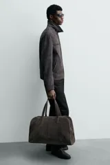 Bolso de viaje de gran tamaño confeccionado en piel con acabado de serraje. Cuenta con un compartimento principal con cierre de cremallera e imanes, interior con bolsillo con cierre y bolsillo plano, y dos asas de mano de piel.