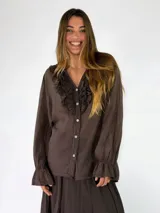Blusa de manga larga color marrón, con cierre frontal de botones, escote en V y detalles de volados fruncidos alrededor del cuello y los puños elásticos.
