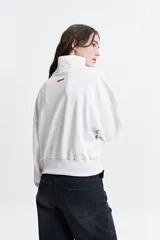 Buzo de felpa color crudo con cuello alto, cierre metálico y bordado de un corazón con rayos en el pecho.