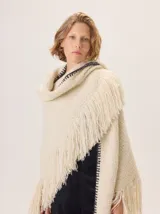 Triángulo de lana color beige con maxi flecos, diseñado por Margara Shaw en colaboración con Manos del Uruguay. Confeccionado con una mezcla de lana merino, mohair, viscosa, algodón y otras fibras, incluyendo lurex y un hilado teñido a mano con lentejuelas.