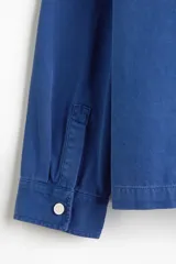 Sobrecamisa de lona de algodón en color azul, con cuello clásico, cierre frontal de botones y tres bolsillos de parche en la parte delantera. Presenta mangas largas con puños abotonados y canesú en la espalda.
