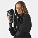 Cartera bandolera negra de charol con tachas plateadas y candado decorativo.