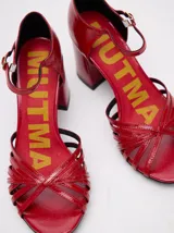 Sandalias de taco alto ancho con diseño retro, confeccionadas en charol negro. Presentan tiras delanteras múltiples entrelazadas, talón cerrado y una correa fina ajustable al tobillo con hebilla metálica.