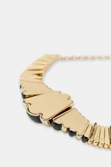Collar tipo babero o gargantilla con cadena dorada y secciones de piezas acrílicas negras y doradas, con forma de tubo acanalado y centro abultado.