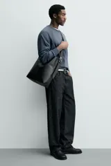 Bolso tipo shopper de cuero sintético negro, con costuras visibles en el frente que dividen el cuerpo en paneles. Presenta un borde superior ondulado y un asa de hombro ajustable.