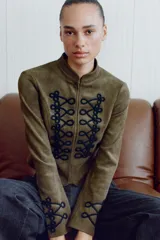 Chaqueta corta de antelina color verde militar, con cuello subido, manga larga y bajo asimétrico. Presenta detalles de alamares negros bordados en contraste en el frente y en los puños.
