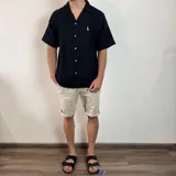 Camisa de manga corta confeccionada en muselina de textura arrugada, con cuello solapa, cierre frontal de botones y pequeño bordado en el pecho.