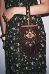Bolso pequeño de mano con asa corta, confeccionado con abalorios negros y diseño floral bordado en tonos rosa y verde. Presenta flecos largos de abalorios en la parte inferior y cierre con botón a presión.