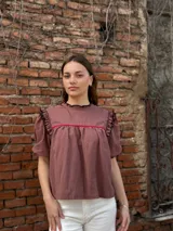 Blusa color chocolate con mangas cortas abullonadas, cuello redondo con volados y detalles de costura en zigzag de color rojo en el pecho y volados oscuros en los hombros.