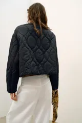 Cazadora corta acolchada de color negro, con cuello redondo, manga larga y cierre frontal con botones. Presenta un diseño de costuras geométricas y bolsillos laterales de vivo.
