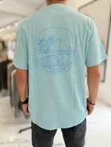 Remera celeste oversize con estampa en la espalda de un paisaje marino con palmeras y olas dentro de un círculo, acompañada de texto.