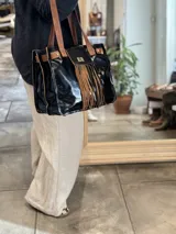 Cartera negra de cuero charol con flecos marrones, doble asa de cuero marrón y cierre con broche metálico.