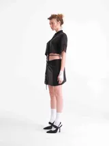 Camisa crop negra de tencel, con mangas cortas y detalles de encaje en el cuello, la botonera frontal, el dobladillo inferior y las mangas. Cuenta con tiras ajustables que se atan alrededor de la cintura.