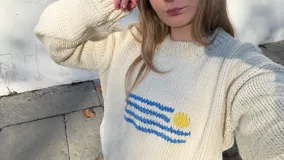 Buzo de punto color crema, tejido a mano en lana merino, con diseño de la bandera de Uruguay bordada en el pecho. Presenta cuello redondo y mangas largas con puños acanalados.