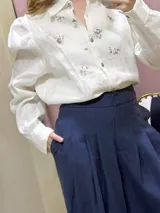 Camisa de lino 100% natural, de color blanco, con mangas largas abullonadas y bordados artesanales a mano en el frente. Cuenta con cuello mao y cierre frontal de botones grises.