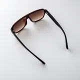 Lentes de sol tipo escudo con armazón de policarbonato marrón oscuro y detalles metálicos dorados en las patillas, con cristales degradados en tonos marrones y protección UV400.
