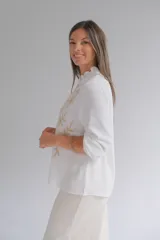 Blusa blanca de corte recto con cuello mao y volado pequeño, manga larga con puños elásticos y cierre frontal de botones. Presenta un delicado bordado floral en color beige en ambos lados del frente.