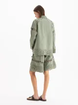 Chaqueta de gabardina liviana color verde militar, con silueta recta, largo por debajo de la cadera y cuello de corderoy. Presenta detalles de encaje en hombros, mangas y ruedo, múltiples bolsillos con solapa y cierre frontal con broches metálicos.