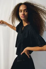 Blusa negra de tejido liviano con corte holgado, manga circular corta y escote en V con tiras finas ajustables. Presenta alforzas verticales en el frente y ojales metálicos decorativos en el escote y los bordes de las mangas.