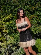 Vestido corto sin mangas, color negro, completamente cubierto de lentejuelas brillantes. Presenta un borde de flecos largos de color rosado en el escote y en el ruedo inferior.
