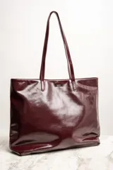 Bolso tipo tote de gran tamaño en color bordó con acabado brillante. Cuenta con doble asa de hombro y placa metálica decorativa con logo en la parte frontal.