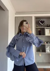 Camisa de corte recto con estampado de cuadros vichy en tonos azul y blanco. Presenta cuello clásico, cierre frontal con botones y volados decorativos que recorren la parte superior de las mangas y los hombros.