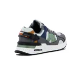 Zapatilla deportiva estilo retro, color gris, blanco, verde y azul marino, con detalles en mostaza y negro. Presenta una capellada con paneles de malla y gamuza, y una suela blanca con salpicaduras negras.