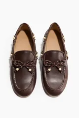 Mocasines estilo náutico de color marrón, con cordones decorativos bicolores y cuentas claras en la parte frontal y laterales. Cuenta con costuras pespunteadas en contraste y suela plana.