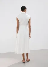 Vestido midi camisero de lino color blanco, sin mangas, con cuello solapa, cierre frontal de botones y cinturón ajustable del mismo género. Cuenta con detalles de costura calada en el frente.