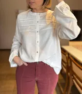 Camisa de lino color blanco con cuello redondo, cierre frontal de botones y detalle de alforzas y encaje en el pecho. Presenta mangas largas con puños abotonados y un corte recto.