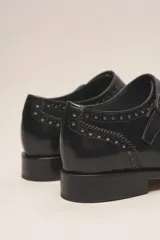 Zapato tipo monk strap con aberturas laterales, confeccionado en cuero vacuno metalizado color bronce. Presenta detalles de perforaciones estilo brogue en la puntera y el talón, y doble hebilla metálica en el empeine.