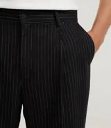 Pantalón de sastrería de corte recto, color negro con finas rayas verticales blancas. Presenta pliegues frontales, cintura ancha con trabillas para cinturón y cierre tradicional.