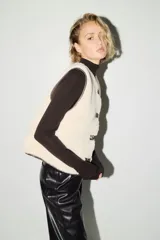 Chaleco de efecto peludito color beige, con cuello redondo, cierre frontal de botones y ribetes en contraste marrón oscuro. Incluye bolsillos laterales discretos y forro interior.