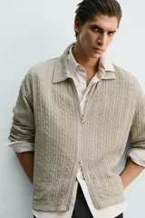 Chaqueta de punto con textura de trenzas verticales, color beige, cuello solapa y cierre frontal con cremallera.