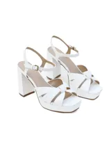 Sandalias blancas de cuero con taco alto de 10 cm y plataforma de 2.5 cm. Cuentan con tiras cruzadas en la parte delantera y cierre de hebilla en el tobillo.